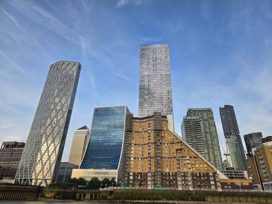 A wide-angle view of Canary Wharf skyline featuring modern high-rise buildings lining the dockside, with a calm waterway reflecting the structures. Visible in the scene are office towers with large gl