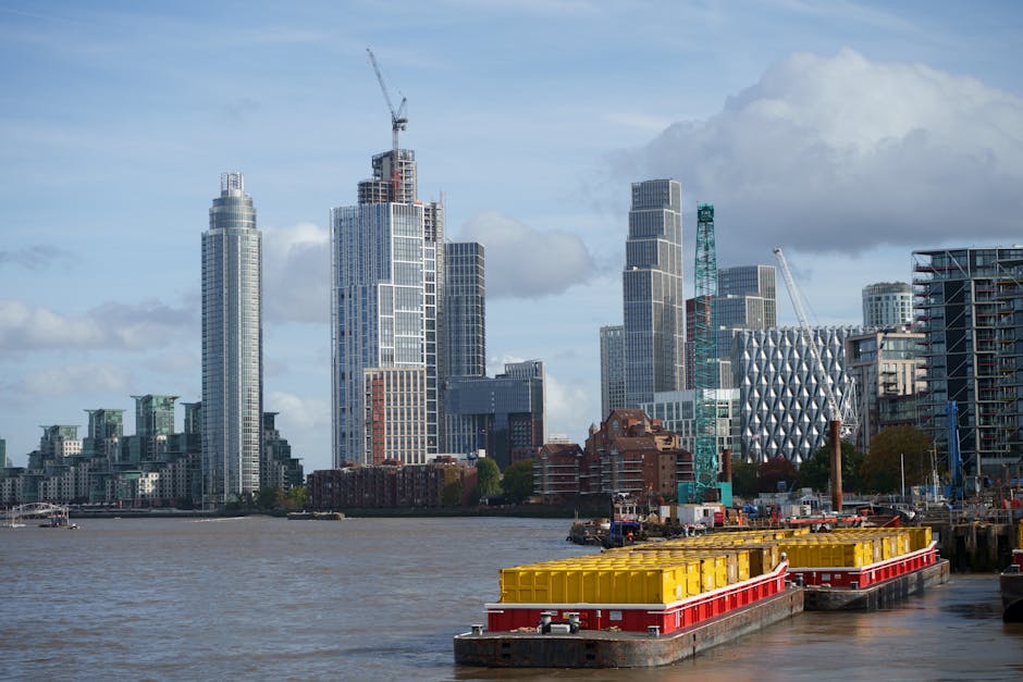 A daytime view of the London skyline from the river, featuring several high-rise buildings under construction with cranes on top, and finished modern office and residential towers with distinctive arc