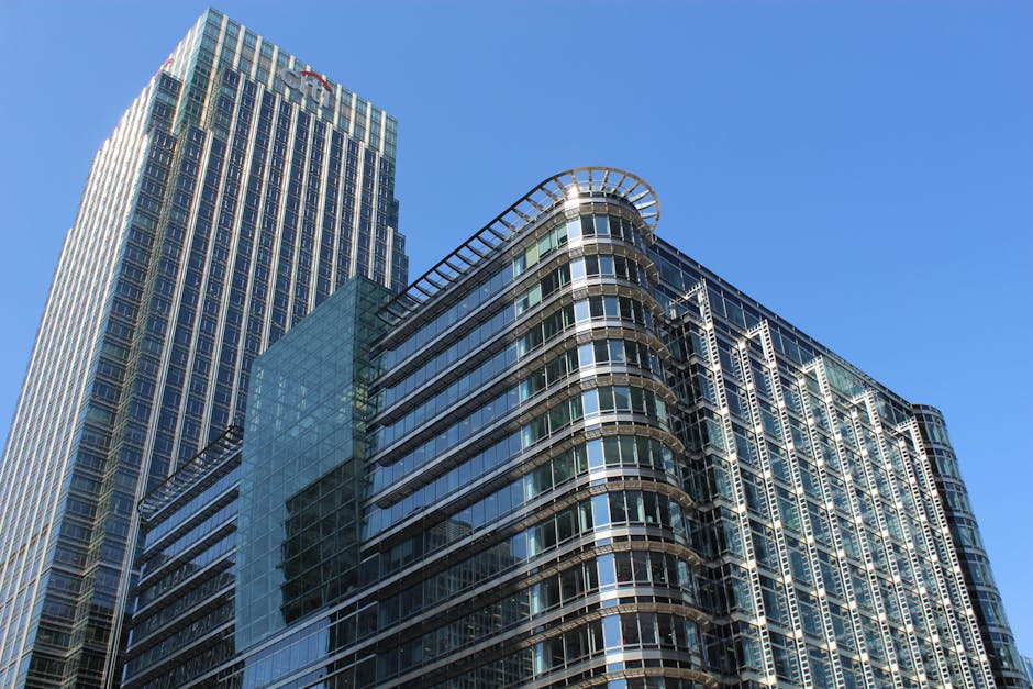 A large floor-to-ceiling glass facade with a grid of vertical and horizontal metal frames, revealing a bustling cityscape of modern office buildings with reflective glass windows and varying architect