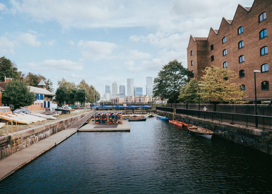 A view of a small marina adjacent to a residential building in Canary Wharf during daytime, with several boats and yachts secured along the water's edge, some covered with tarps and others with wooden